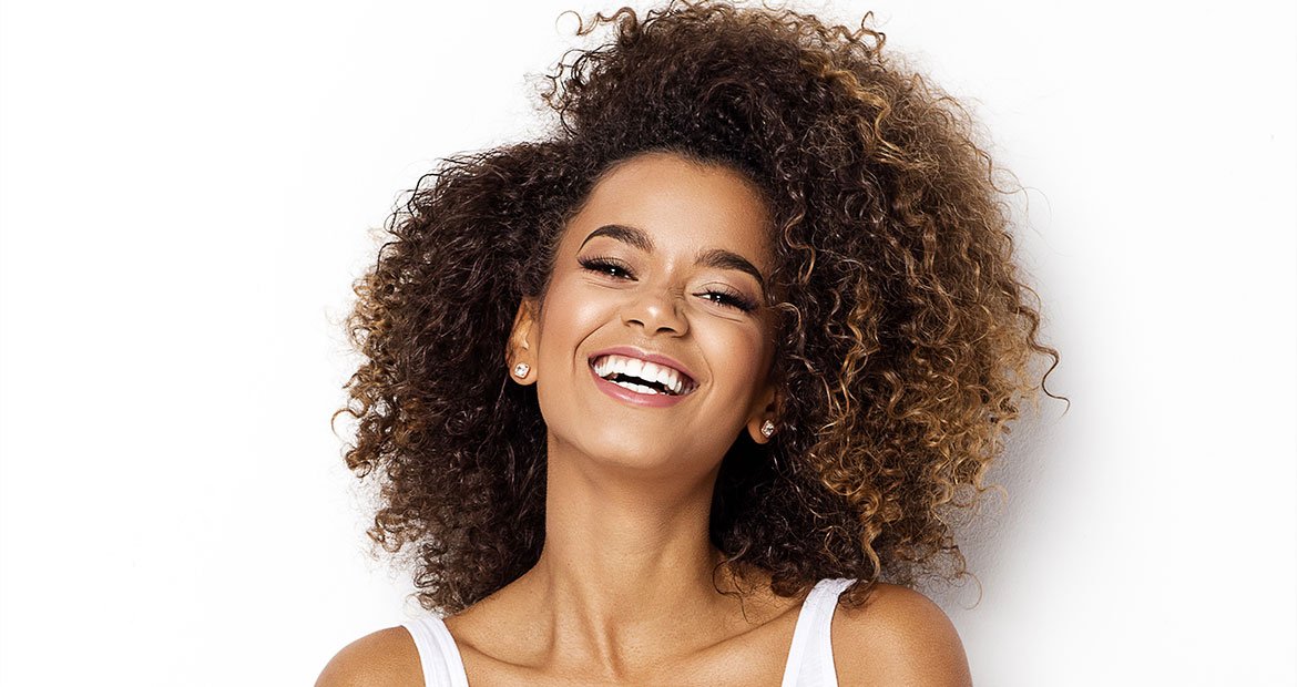 Morena iluminada com cabelo cacheado curto e volumoso, mostrando um sorriso alegre
